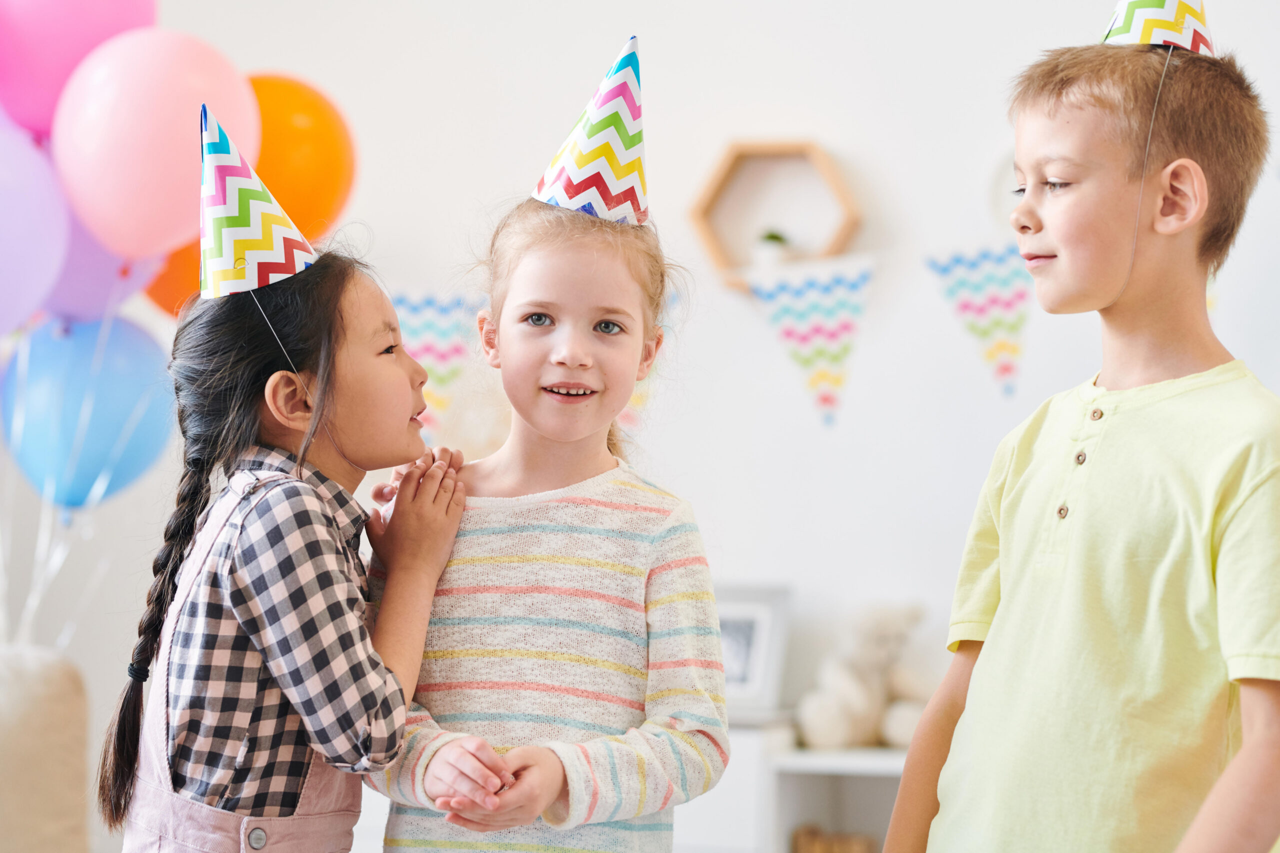 Cute little girl whispering something to one of her friends at home birthday party during game after festive dinner